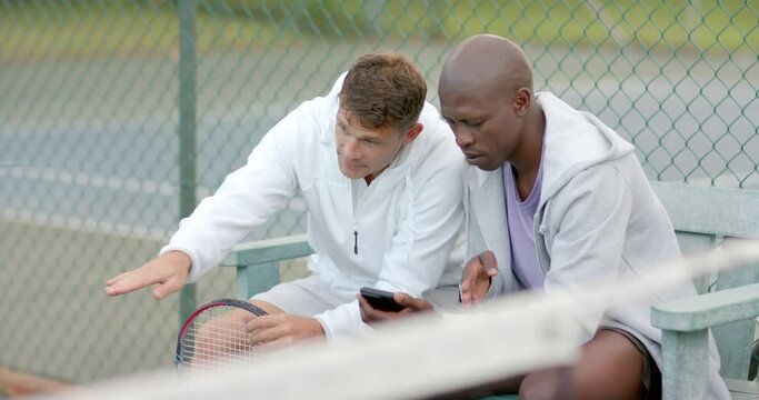 Happy diverse male friends looking at smartphone sitting on bench at tennis court, slow motion