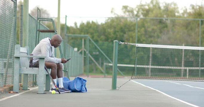Happy african american man using smartphone sitting on bench at tennis court, slow motion