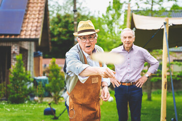 Belgian senior men engaged in a traditional disc-throwing game (Bakschieten) in their country house backyard, one throwing a small disk, the other observing with hands on hips.