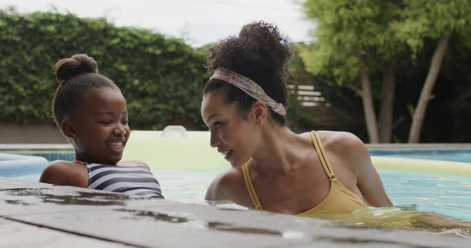 Happy african american mother and daughter playing in swimming pool in garden, slow motion