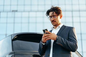 Young businessman standing near luxury car and using smartphone