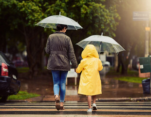 Back, raincoat or umbrella with a mother and daughter walking across a street in the city during winter. Autumn, crosswalk or park with a woman and female child holding hands while crossing a road © Arnell K/peopleimages.com