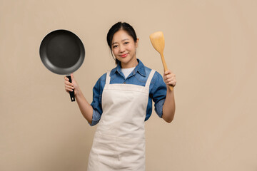 Young Asian woman housewife wearing kitchen apron cooking and holding pan and spatula isolated on...