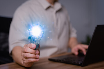 Adult man holding a light bulb while working on a computer laptop to think and generate new ideas through technological innovation and creativity. Idea inspired by online technology and innovation.