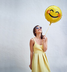 Woman, balloon and happy by wall in with emoji, smile and face for fashion sunglasses at party,...