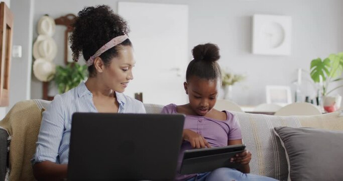 Happy african american mother and daughter sitting on sofa, using laptop and tablet, slow motion