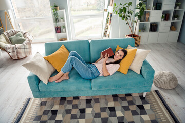 Photo of positive cheerful girl lying on comfy divan reading paper book in modern house interior flat