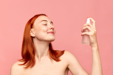 Portrait with redhead girl, model using moisturising spray on face with closed eyes over pink studio background. Facial cosmetics product, masks