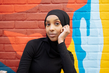 Portrait of young woman wearing hijab sitting against mural