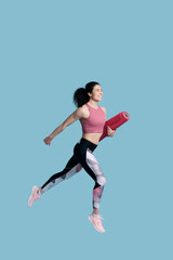 Fototapeta na wymiar Smiling athletic woman in sportswear carrying yoga or fitness mat while running over blue background