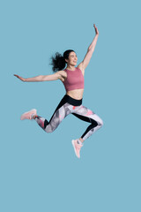 Fototapeta na wymiar Full size young athletic female fitness trainer doing gym exercises, jumping high on blue background