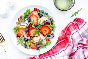 Summer strawberry and grilled chicken healthy salad with arugula, blueberries, red onion and walnuts, white table. Fresh useful dish for healthy eating