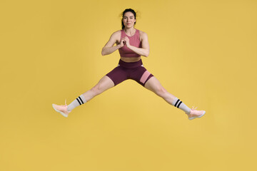 Fototapeta na wymiar Young sportswoman performing stretching exercises and jumping high up, on isolated yellow background