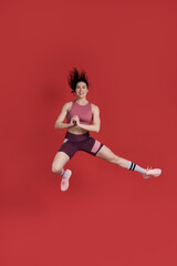Fototapeta na wymiar Female athlete in activewear, smiles at camera, performs stretching exercise, jumps over red walll
