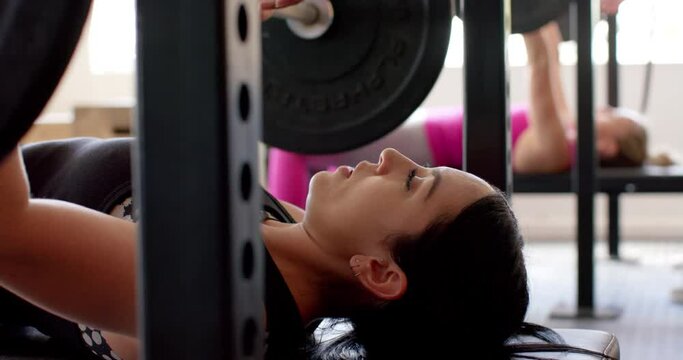 Determined unaltered biracial woman weight training at gym bench pressing barbell, in slow motion