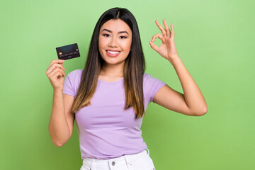 Photo of young optimistic woman wear purple t-shirt hold plastic card show okey sign enjoy convenient payment isolated on green background