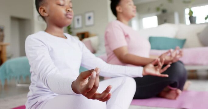 Happy unaltered african american mother and daughter doing yoga meditation at home, in slow motion