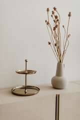 A golden plated cake stand and a beige ceramic vase with dried flowers on the cabinet. Copy space, white background. Modern home decoration.