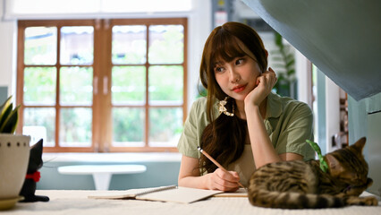 A beautiful dreamy Asian girl is writing her diary while her cute cat is on a table