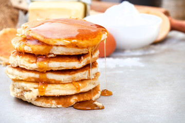 Fresh Pancakes stacked on kitchen table with ingredients in background and with syrup dripping down