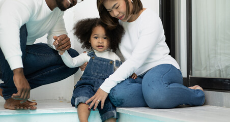 Happy multiracial family having fun at home together. Portrait of multiethnic father mother and little biracial daughter sitting at swimming pool relaxing. Diverse ethnic dad and mom playing with kid. - Powered by Adobe