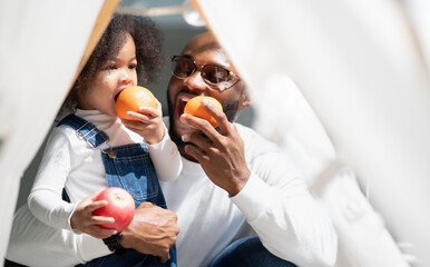 Happy multiracial family having fun at home together. Portrait of multiethnic father and little biracial daughter playing with apple at backyard camping. Diverse ethnic dad laughing with cheerful kid.