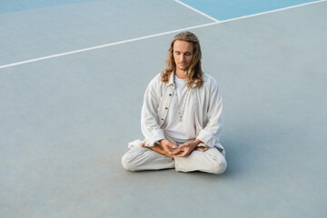 long haired man in white cotton clothes practicing yoga in lotus pose on outdoor stadium