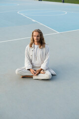 full length of stylish man with long fair hair meditating in easy yoga pose on stadium outdoors