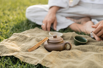 Fototapeta na wymiar partial view of yoga man sitting near linen rug with palo santo stick and clay teapot with cups on green lawn