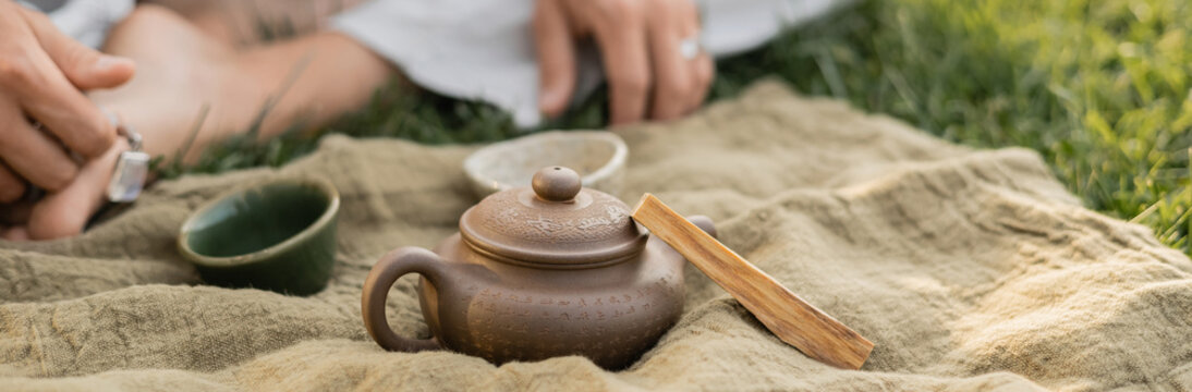 partial view of blurred man sitting on green lawn near linen rug with clay teapot and cups, banner - Powered by Adobe