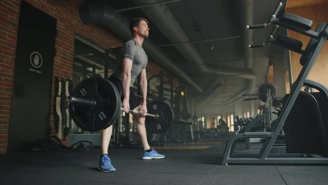 Young handsome athletic man lifts barbell in spacious gym with strength training equipment in black colors. Sportsman performs deadlift, legs far apart. High quality 4k footage