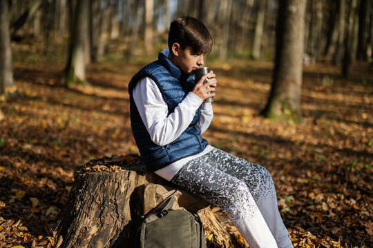 Autumn outdoor portrait of teenager boy sit in stump and drink tea from thermos cup in forest.
