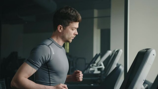 Medium close up profile shot of handsome athletic man running on treadmill in gym. Man is looking on display. Row of treadmills in the background. High quality 4k footage