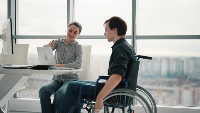 Young male office worker in a wheelchair talking to female colleague while showing her ideas using laptop in co-working space. Disability and handicap concept. Office modern people at work.