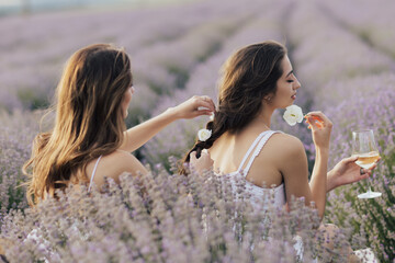 Two sisters enjoying a picnic with fresh fruit and wine while sitting and chatting on field of lavender. One girl braids another girl's hair.	