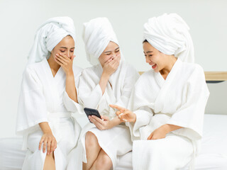 Millennial three Asian female customers friends in white clean bathrobes and towels have appointment at massage resort sitting on chair holding smartphone taking selfie photo together after massaging