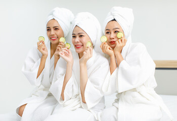 Millennial three Asian female customers friends in white clean bathrobes and towels have appointment at massage resort sit on chair holding sliced cucumbers placed on closed eyes treatment together