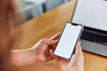 A woman holding a mobile phone with an empty white screen, sitting in front of the laptop.