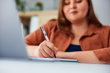 A plus-size woman writes notes while working on the laptop at the table.