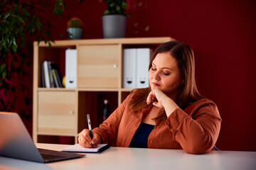 A curvy businesswoman writing notes in a notebook and working on a laptop.