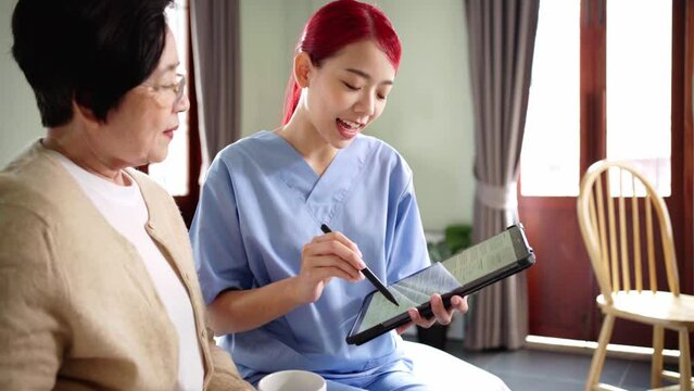 Asian woman nurse wearing scrubs report the health status of Senior Asian woman with tablet in the bedroom. Caregiver visit at home. Home health care and nursing home concept.