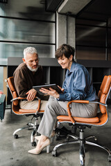 Cheerful mature colleagues discussing work projects on tablet in office