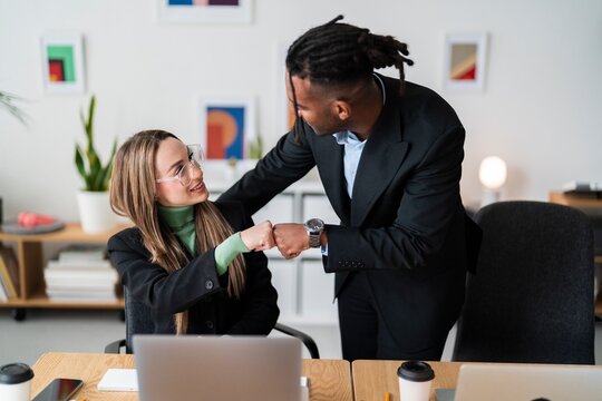 Positive young multiracial coworkers in formal outfits smiling and looking at each other while fist bumping during successful meeting in modern office