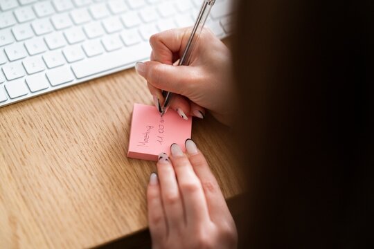 Unrecognizable female entrepreneur with manicure writing information about business meeting on sticky notepad while sitting at wooden table near keyboard in modern office