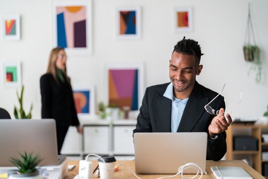 Smiling ethnic businessman in formal clothes sitting at wooden desk with coffee in paper cup and using laptop during workday in light modern office