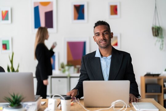 Smiling ethnic businessman in formal clothes sitting at wooden desk with coffee in paper cup and using laptop during workday in light modern office