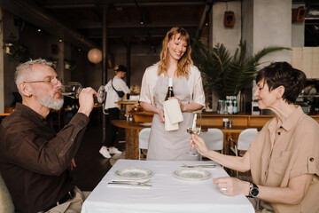Positive young blonde waitress looks at old couple drinking wine in cafe
