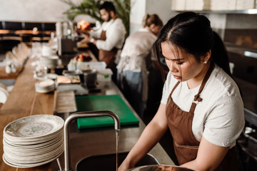 Confident asian female chef in apron washes dishes behind counter in cafe 