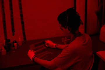 Young photographer working with photo film at table in darkroom with red light