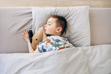 Happy cute tired little boy lies on comfort bed with closed eyes, hugs toy dog, sleeping in bedroom interior, top view, free space, close up. Health care, childhood, rest, relax at night